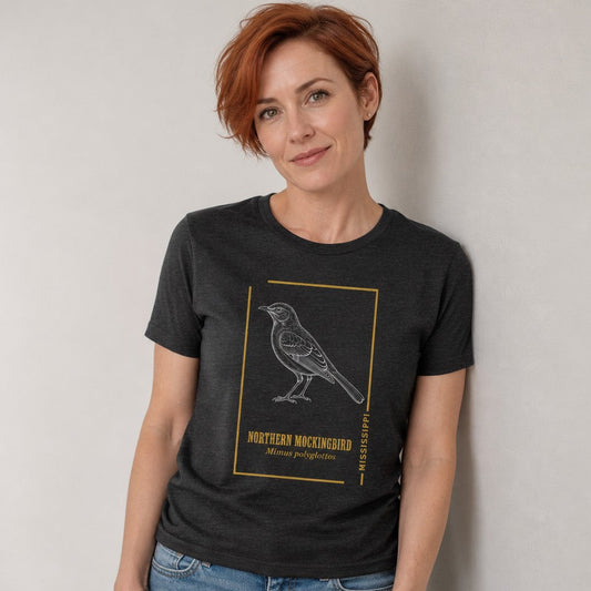 Woman wearing a dark grey heather Bella Canvas 3001 t-shirt featuring a vintage hand-sketched Northern Mockingbird illustration with botanical gold accents, vintage naturalist style, for birdwatchers and classic wildlife lovers, by Crossroads Dandelion