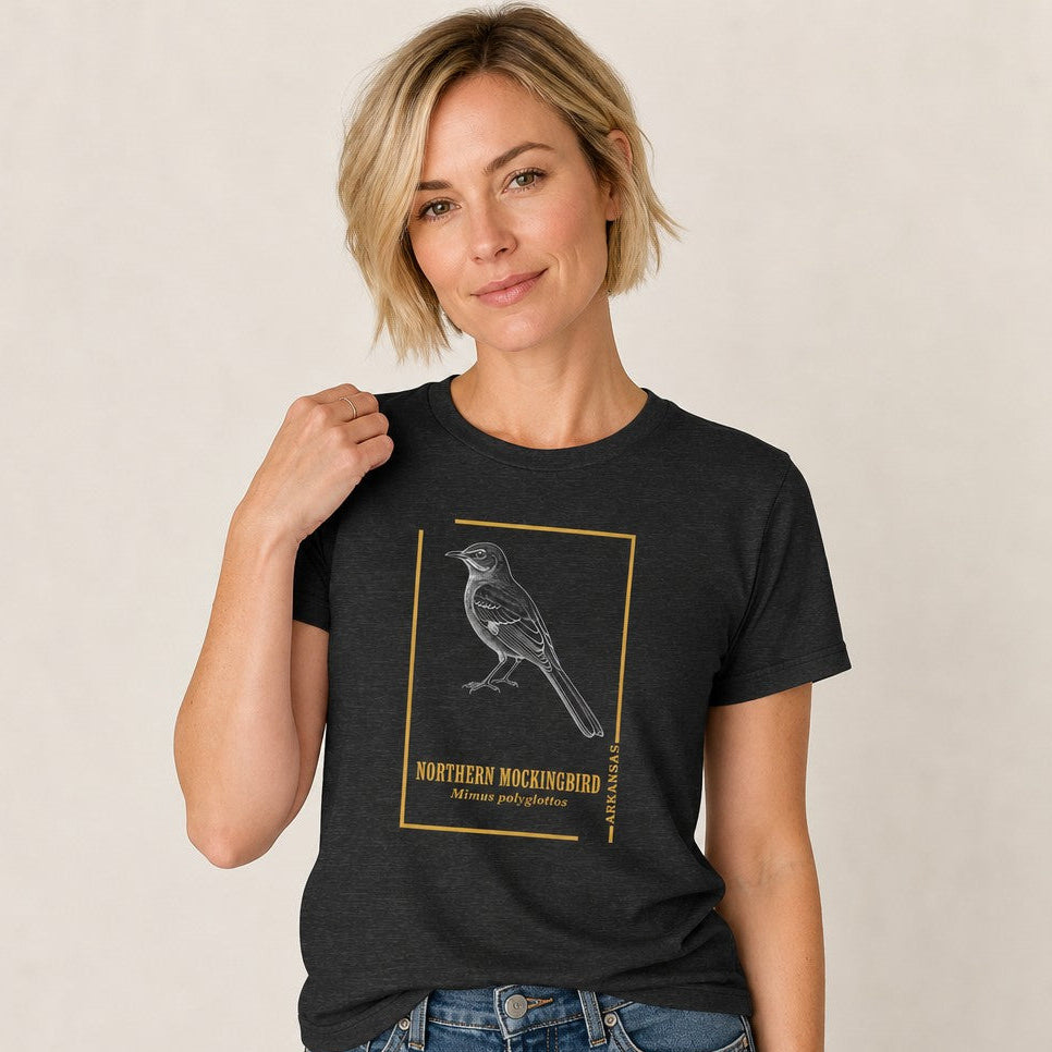 Woman wearing a dark grey heather Bella Canvas 3001 t-shirt with a vintage-style line art illustration of a Northern Mockingbird and gold border, inspired by antique field guides, perfect for birdwatchers and nature lovers, by Crossroads Dandelion