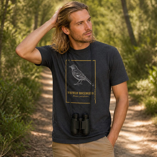 Man wearing a dark grey heather Bella Canvas t-shirt with vintage line art of a northern mockingbird in white ink, framed in gold, standing on a forest trail with binoculars, perfect for birdwatchers and botanical enthusiasts, by Crossroads Dandelion