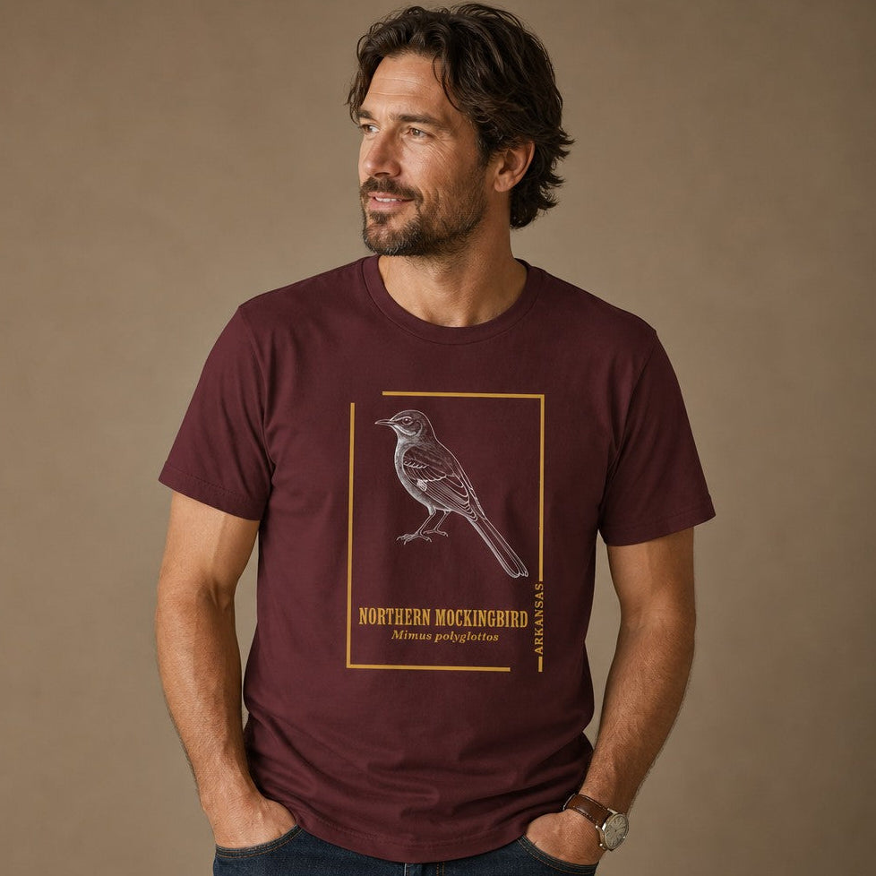 Man wearing a maroon Bella Canvas 3001 t-shirt featuring a vintage line art illustration of a Northern Mockingbird within a gold rectangular border, styled like an antique field guide for birdwatchers and nature enthusiasts, casual indoor scene, for men and bird lovers, by Crossroads Dandelion