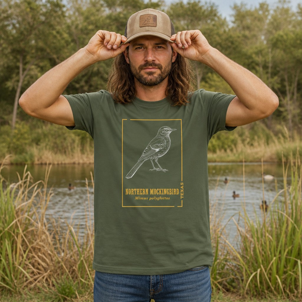Man wearing a military green Bella Canvas 3001 t-shirt with a hand-drawn, vintage-style northern mockingbird illustration in white line art, accented with gold botanical framing and classic serif text, standing by a pond in a natural outdoor setting, perfect for birdwatchers and Texas natives, by Crossroads Dandelion