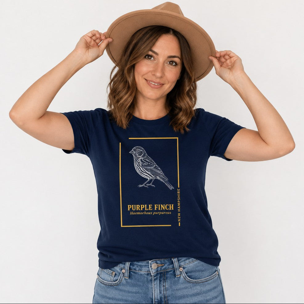 Woman in navy Bella Canvas 3001 t-shirt featuring a vintage hand-illustrated Purple Finch in detailed white ink, framed by golden lines with elegant serif typography, evoking a botanical field guide style for wildlife and New England nostalgia lovers, by Crossroads Dandelion