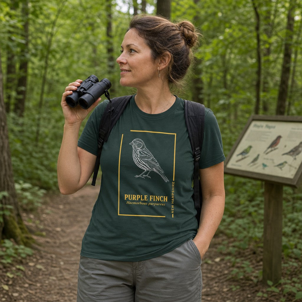 Woman wearing a forest green Bella Canvas 3001 t-shirt featuring a vintage-style hand-illustrated Purple Finch in white ink, framed with golden lines and botanical field guide typography, hiking in a lush green forest, perfect for wildlife lovers and birdwatchers, by Crossroads Dandelion