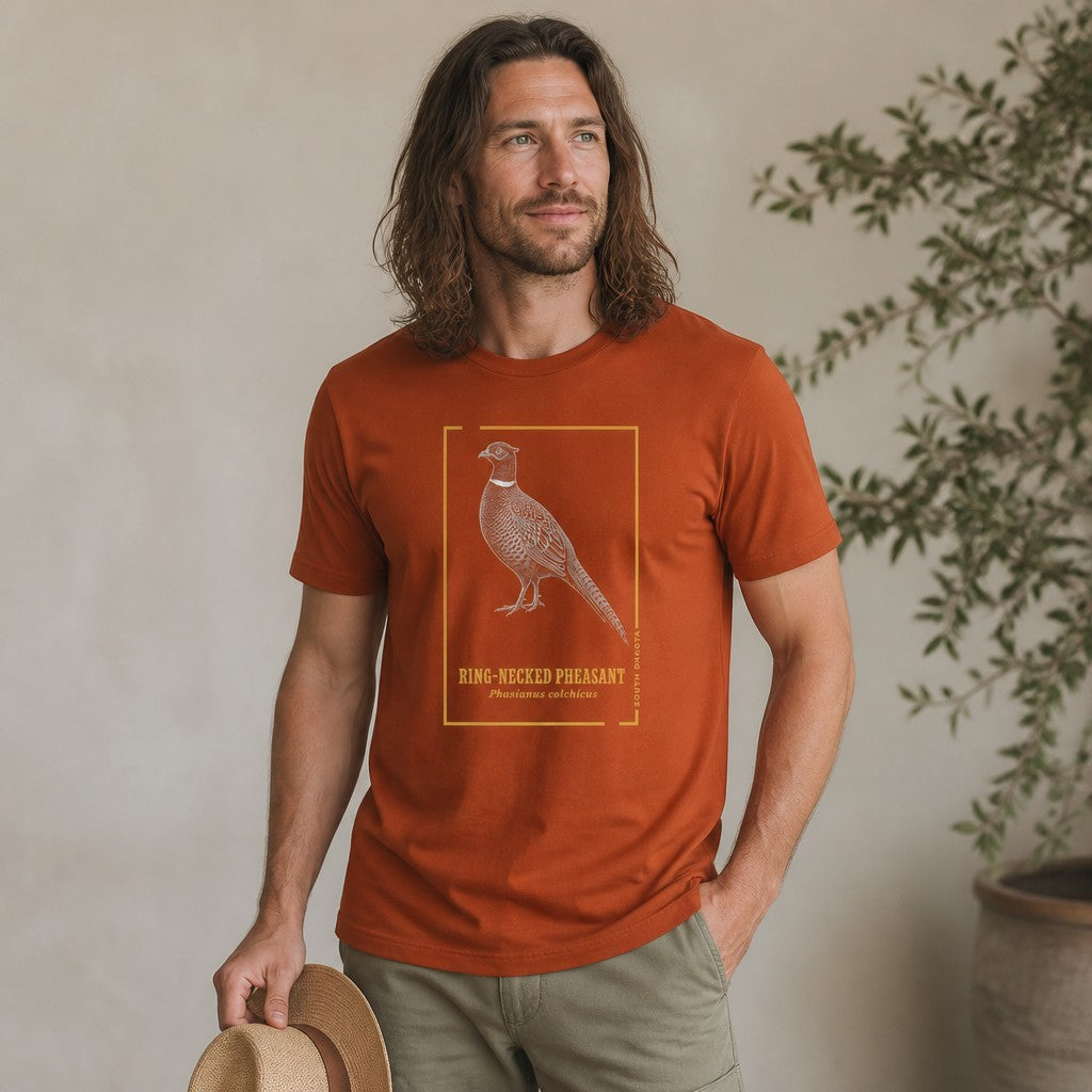 Man wearing autumn orange Bella Canvas 3001 t-shirt featuring a vintage hand-illustrated ring-necked pheasant with gold botanical accents and elegant block lettering, perfect for birdwatchers, nature lovers, and state symbol enthusiasts, casual indoor botanical setting, by Crossroads Dandelion