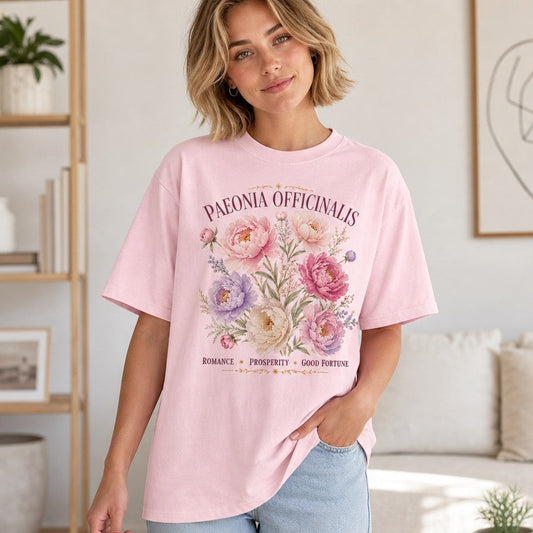 Woman wearing a Comfort Colors 1717 t-shirt in blossom pink featuring a vintage watercolor paeonia officinalis botanical print with celestial accents, feminine and enchanting cottagecore style, perfect for floral lovers in a cozy living room, by Crossroads Dandelion