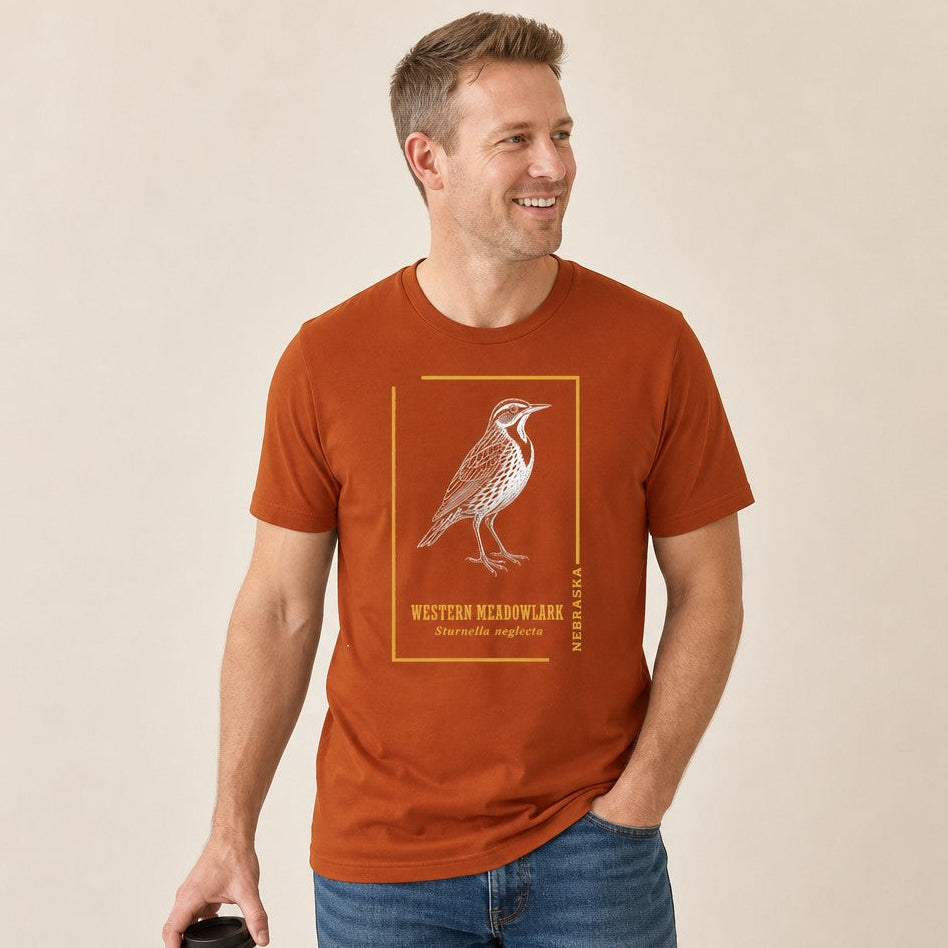Man wearing a Bella Canvas 3001 t-shirt in autumn orange featuring a vintage hand-drawn Western Meadowlark white ink design with gold frame and botanical typography, classic field guide style for birders and artistic nature lovers, by Crossroads Dandelion