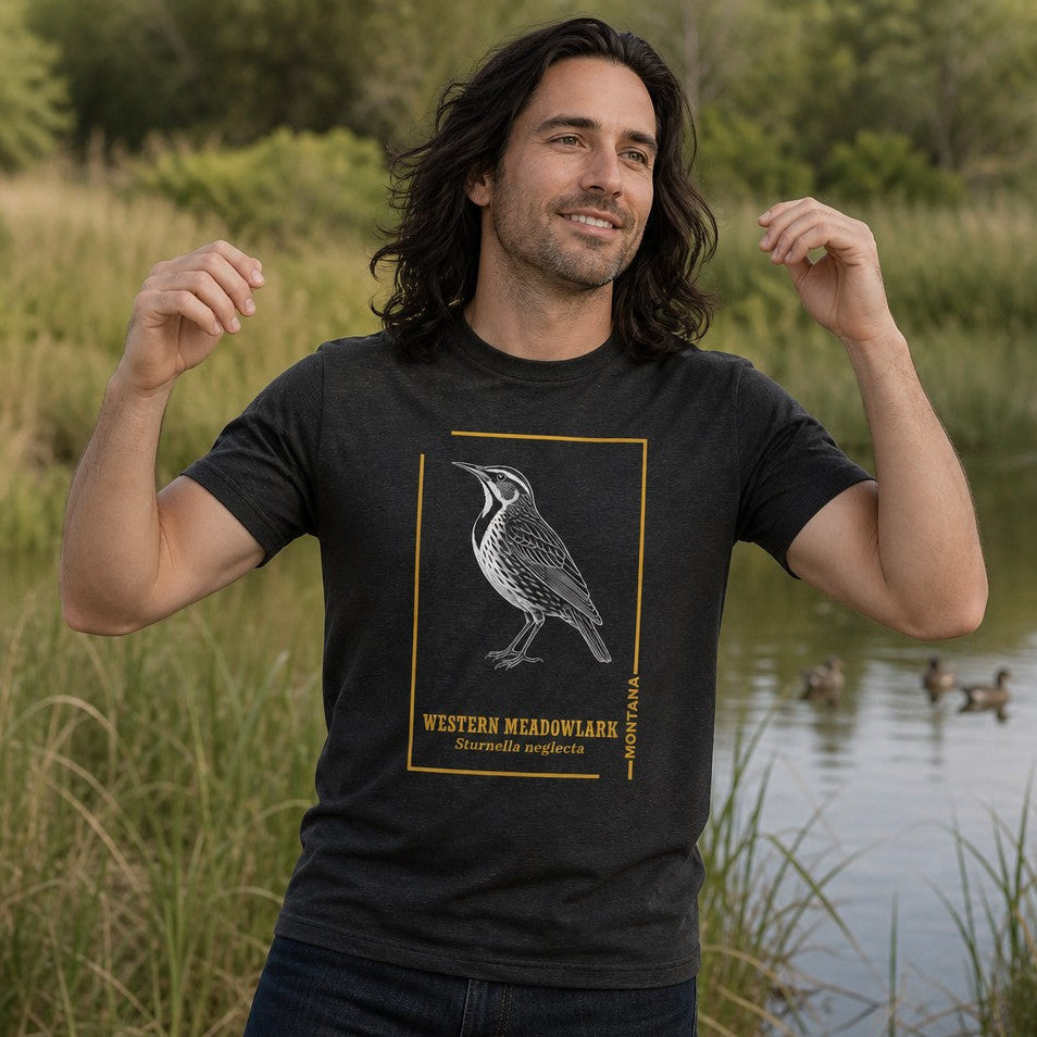 Man wearing a dark grey heather Bella Canvas 3001 t-shirt featuring a hand-drawn vintage etching of a Western Meadowlark framed in gold lines and elegant typography, standing by a pond with reeds and ducks, ideal for birdwatchers, classic nature art lovers, and outdoor heritage style enthusiasts, by Crossroads Dandelion