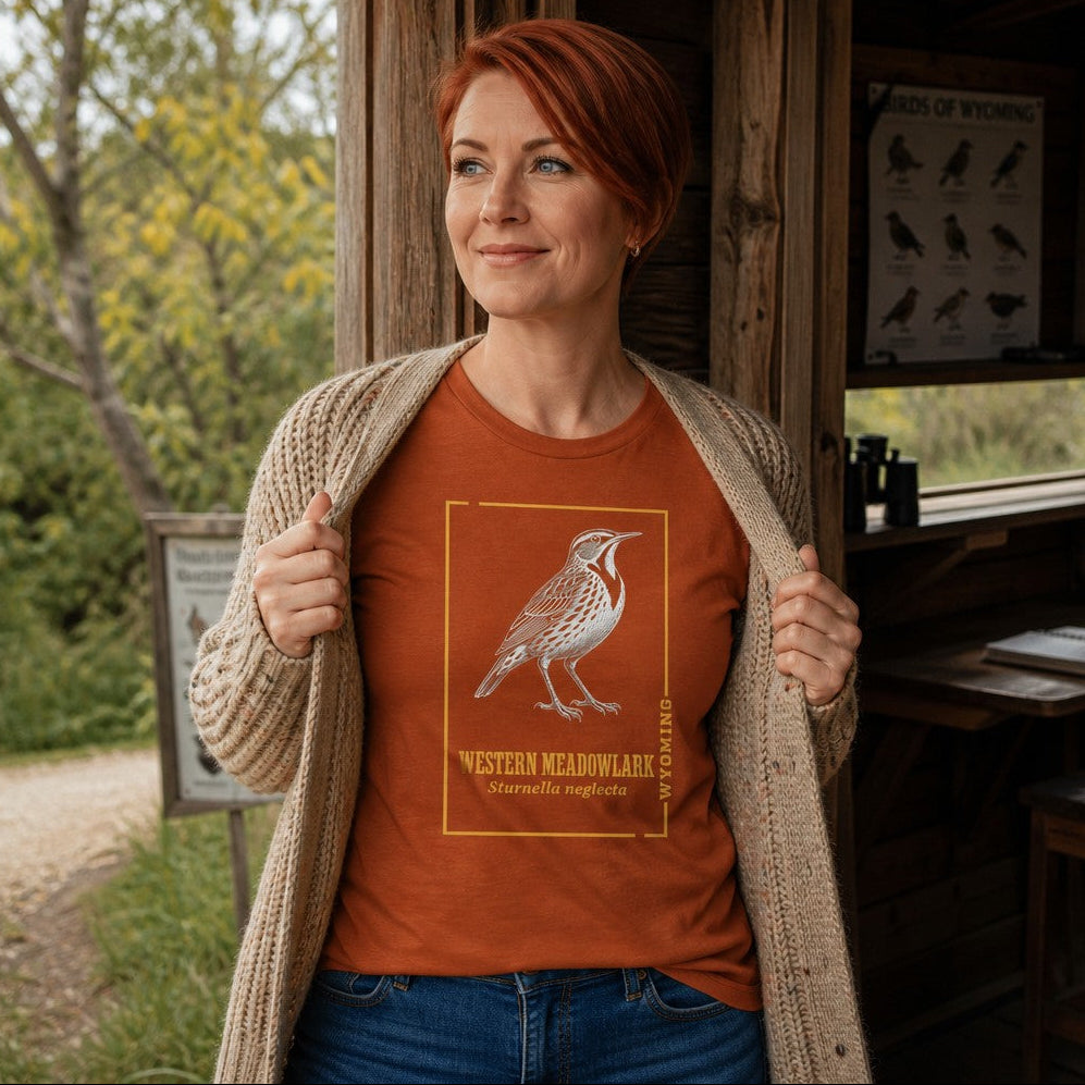 Woman with short red hair wearing an autumn-colored Bella Canvas 3001 t-shirt featuring a vintage hand-inked Western Meadowlark design bordered in gold, inspired by classic field guide and nature journal illustrations, perfect for birdwatchers and Wyoming enthusiasts, standing in a rustic birdwatching cabin, by Crossroads Dandelion
