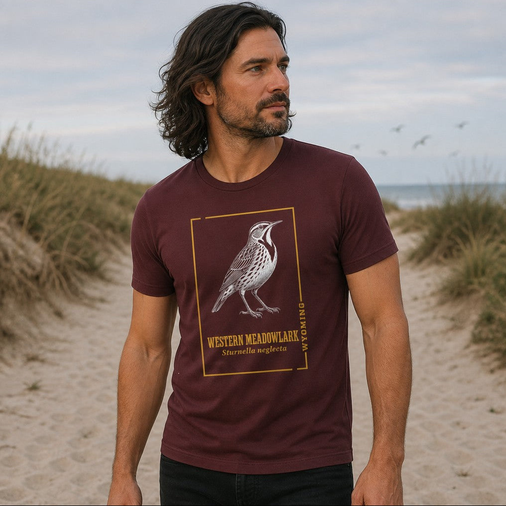 Man wearing a maroon Bella Canvas 3001 t-shirt with a vintage hand-inked illustration of the Western Meadowlark framed in gold, evoking classic field guide style, ideal for birdwatchers and Wyoming nature lovers, standing on a coastal sandy path, by Crossroads Dandelion