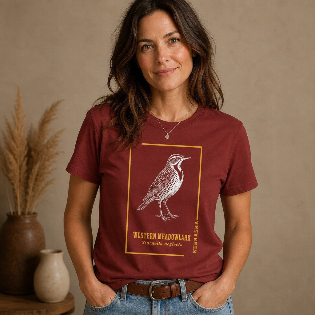 Woman wearing a cardinal red Bella Canvas 3001 t-shirt with a vintage-inspired white hand-drawn Western Meadowlark illustration, golden botanical accents, perfect for birders and nature lovers, cozy indoor setting, by Crossroads Dandelion