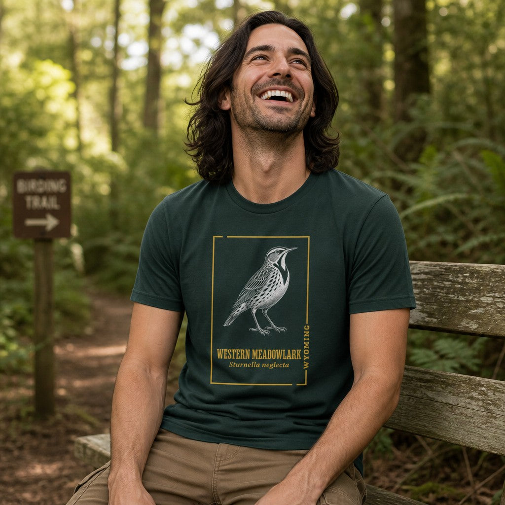 Man wearing a forest green Bella Canvas 3001 t-shirt featuring a vintage hand-inked illustration of the Western Meadowlark within a golden border, perfect for birdwatchers and Wyoming nature lovers, pictured sitting on a bench along a woodland trail, by Crossroads Dandelion