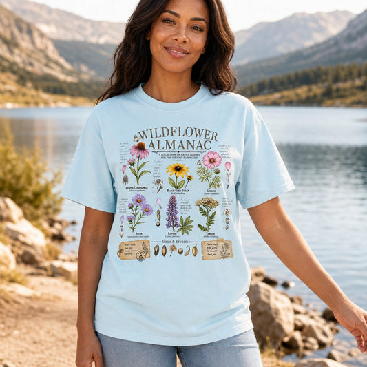 Woman wearing chambray Comfort Colors 1717 t-shirt with vintage watercolor wildflower almanac illustration, botanical notes, seeds, butterflies, and journal-style ephemera, lakeside in the mountains, designed for creative nature-loving women, by Crossroads Dandelion