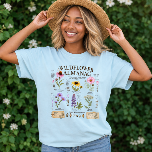 Smiling woman in chambray blue Comfort Colors t-shirt with vintage botanical wildflower almanac illustration featuring hand-drawn native flowers, butterflies, and cursive notes in earthy tones, outdoors in a garden, educational nature lover gift, by Crossroads Dandelion