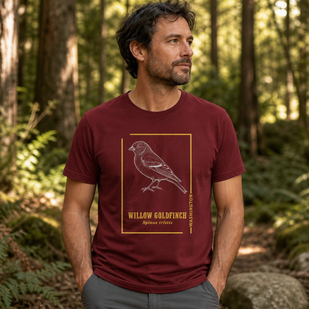 Man wearing a cardinal red Bella Canvas 3001 t-shirt with a vintage etching style willow goldfinch framed in gold, inspired by field journal sketches, photographed in a lush forest setting; ideal for birdwatchers, nature lovers, and botanical art enthusiasts, by Crossroads Dandelion