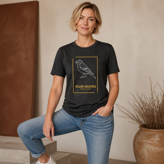 Woman in dark grey heather Bella Canvas 3001 t-shirt featuring a vintage etching willow goldfinch framed in gold accents and serif text, field journal style, cozy indoor setting, ideal for nature lovers and birdwatchers, by Crossroads Dandelion
