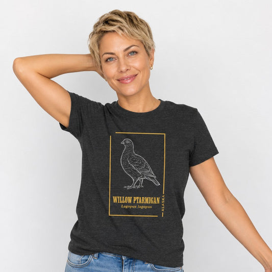 Woman wearing a dark grey heather Bella Canvas t-shirt featuring a hand-drawn vintage illustration of the willow ptarmigan, Alaska’s state bird, accented with golden botanical framing, cottagecore field journal style for birdwatchers, wildlife enthusiasts, and nature lovers, by Crossroads Dandelion
