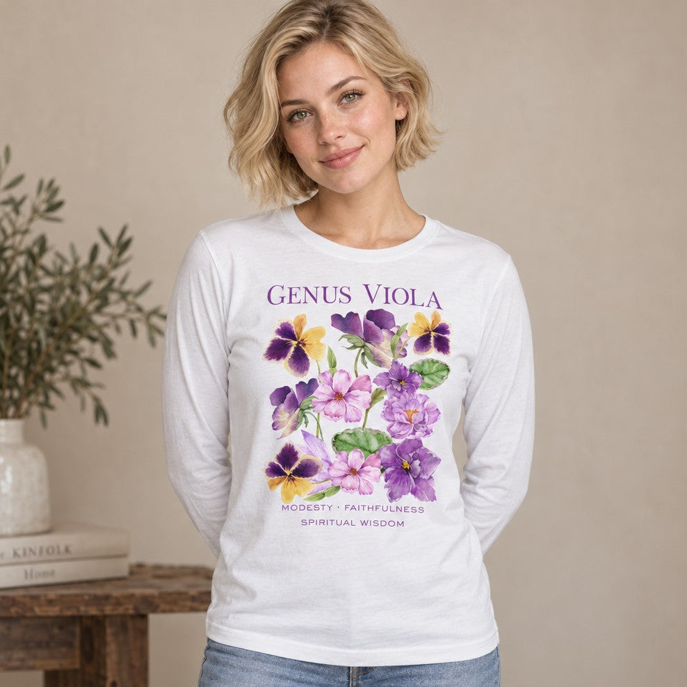 Woman wearing a white Bella Canvas 3501 long sleeve shirt featuring a delicate watercolor illustration of purple, lavender, and yellow viola flowers with soft green leaves, elegant serif 'Genus Viola' text, and affirmations 'Modesty, Faithfulness, Spiritual Wisdom'; serene botanical design for nature and floral lovers, by Crossroads Dandelion