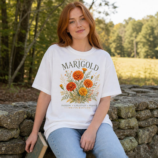 Woman wearing white Comfort Colors 1717 t-shirt with watercolor marigold bouquet design, intricate orange and yellow blooms with botanical foliage, cottagecore floral tee for artists, gardeners, flower lovers, photographed outdoors on a stone wall, by Crossroads Dandelion