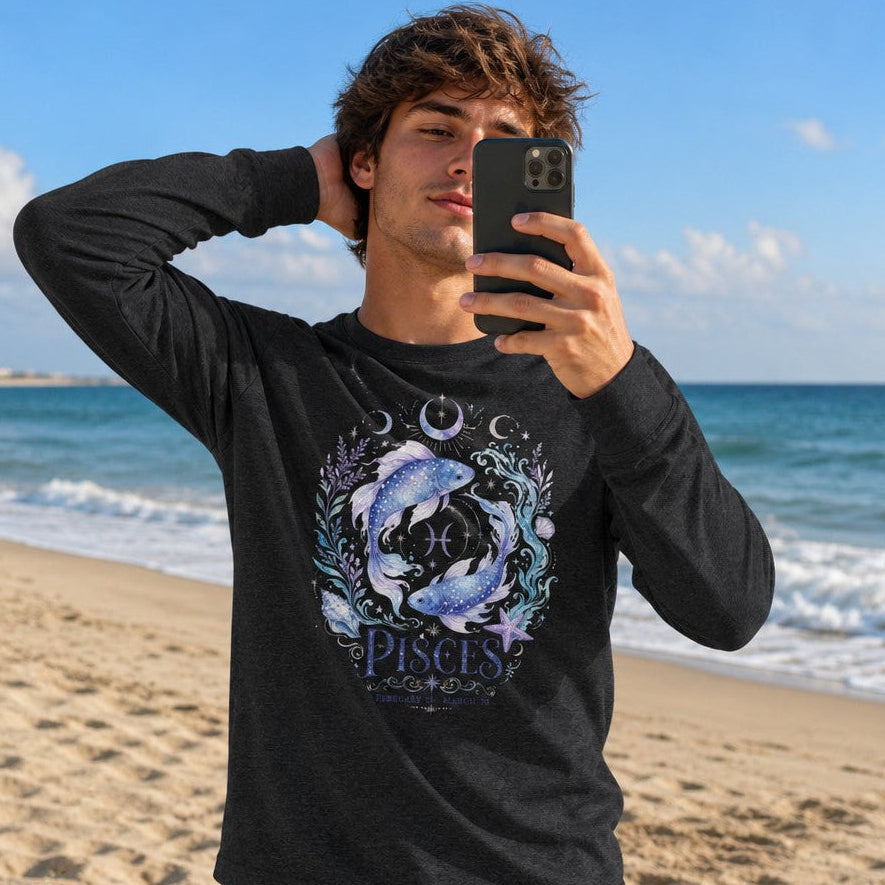 Man wearing a dark grey heather Bella Canvas 3501 long sleeve shirt with a watercolor Pisces design featuring luminous koi fish, violet florals, celestial moons, and sea foliage, standing on a sunny beach, perfect for astrology lovers and gentle souls, by Crossroads Dandelion