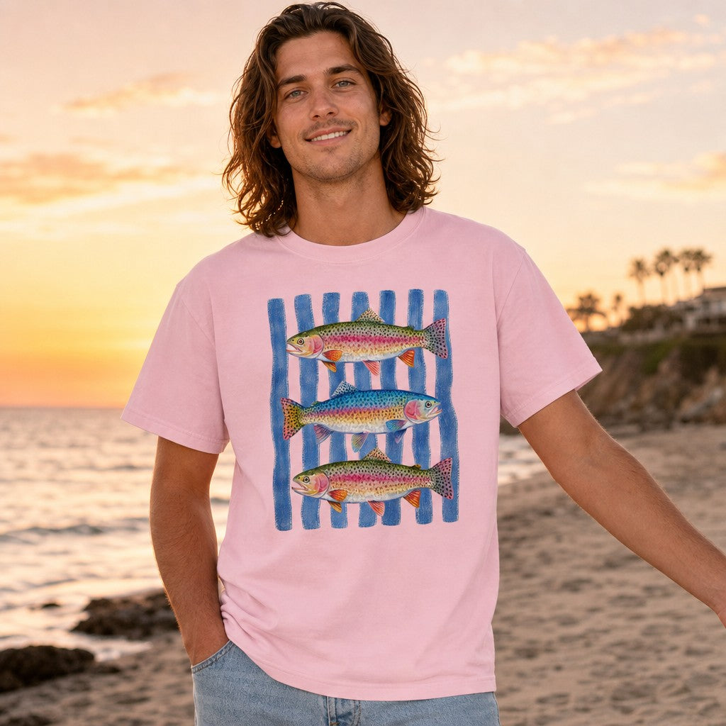 Man wearing a blossom pink Comfort Colors t-shirt featuring a hand-painted watercolor trio of rainbow trout on rippling blue stripes, standing on a beach at sunset, coastal style for nature and fishing enthusiasts, by Crossroads Dandelion