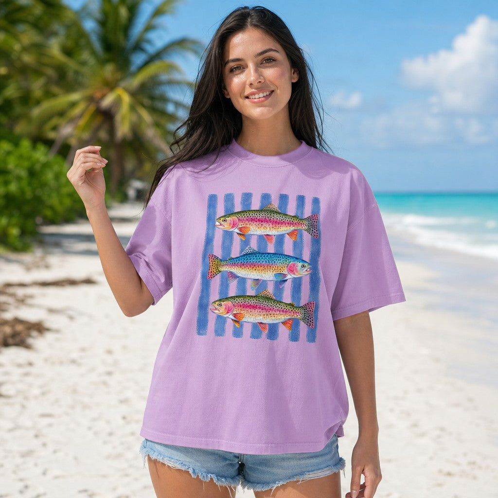 Woman in orchid Comfort Colors 1717 t-shirt featuring a hand-painted watercolor trio of rainbow trout over denim blue rippling stripes, standing on a tropical beach, perfect for nature lovers, fishing fans, and outdoor enthusiasts, by Crossroads Dandelion