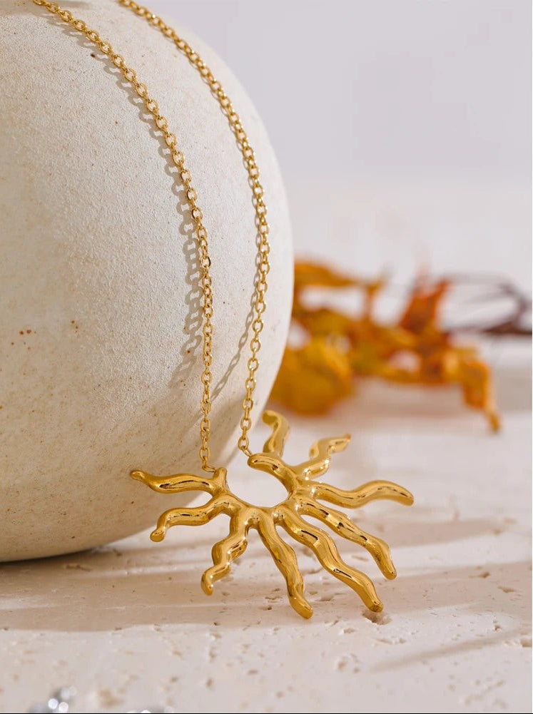 Gold wavy sun pendant necklace draped over a cream stone with autumn leaves behind