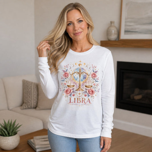 Woman in a white Bella Canvas 3501 long sleeve shirt featuring a whimsical watercolor Libra design with golden scales, pink roses, celestial moon backdrop, floral botanical details, poetic and ethereal, in a cozy living room setting, zodiac and nature art lovers, by Crossroads Dandelion