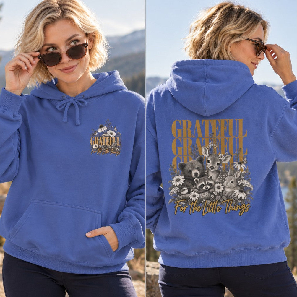 Woman wearing a flo blue hoodie featuring a whimsical woodland animals and floral design with 'Grateful For The Little Things' text, set outdoors in a mountain scene, hoodie aimed at women and nature lovers, by Crossroads Dandelion