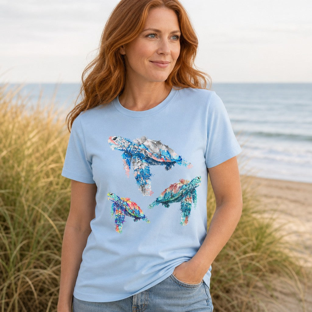 Woman wearing a baby blue Bella Canvas 3001 t-shirt featuring a dreamy watercolor illustration of three sea turtles in blue, teal, and coral hues, standing by the beach with ocean in the background, by Crossroads Dandelion