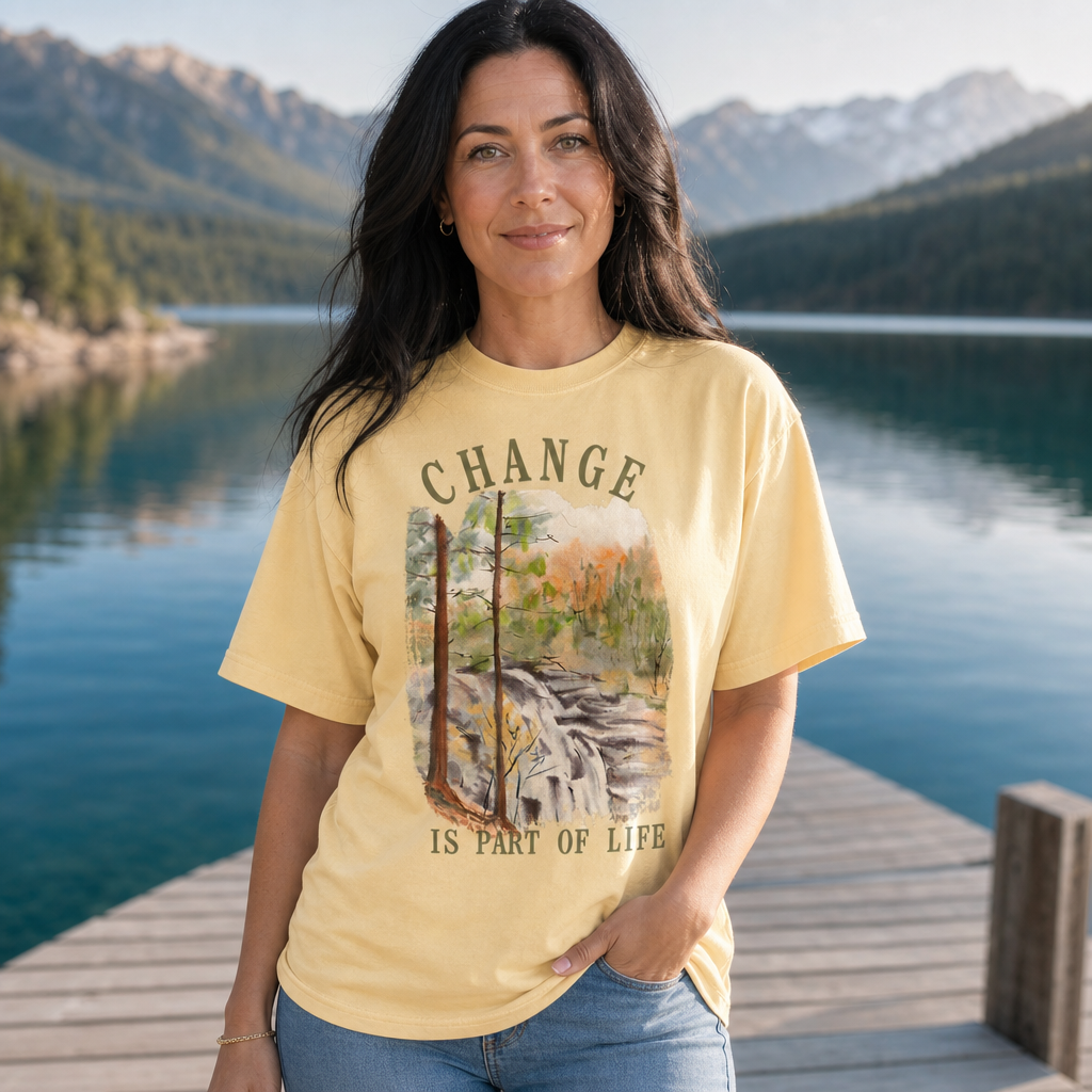 Woman wearing a butter yellow Comfort Colors 1717 t-shirt featuring a watercolor-style forest and river scene with the motivational phrase 'Change is part of life' in bold lettering, standing on a dock by a scenic lake and mountains, by Crossroads Dandelion