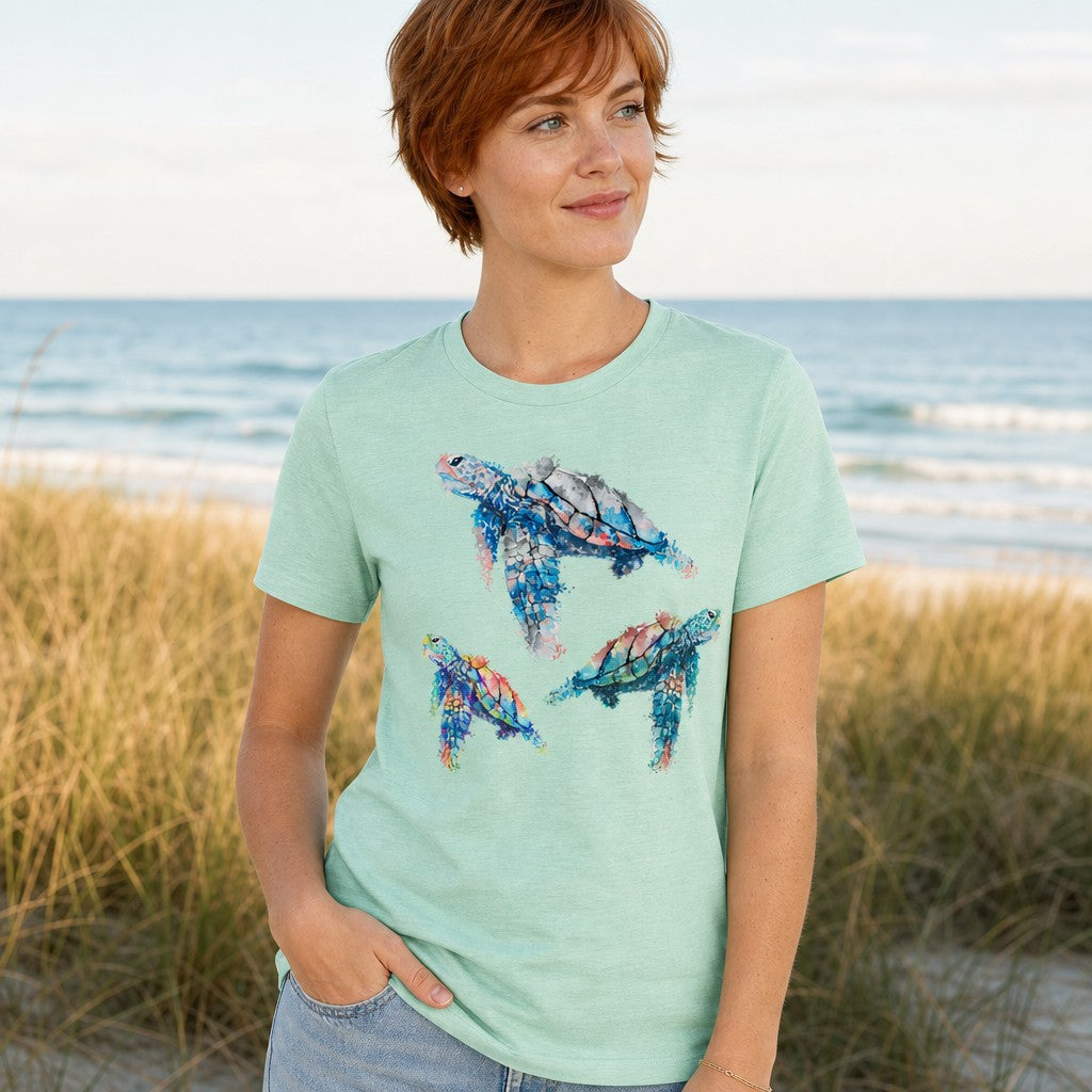 Woman wearing a heather mint Bella Canvas 3001 t-shirt with a dreamy watercolor sea turtle illustration, standing on a beach with ocean in the background, by Crossroads Dandelion