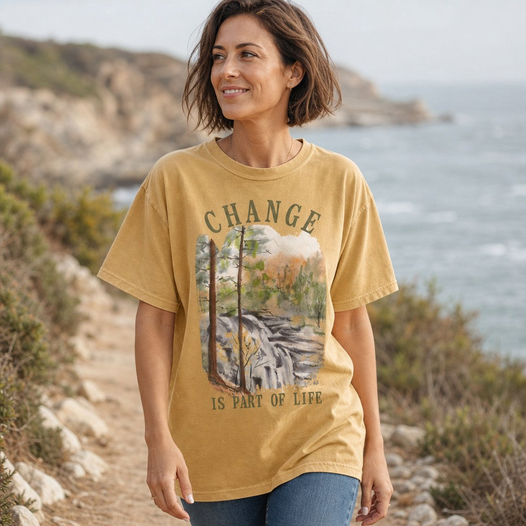 Woman wearing a mustard Comfort Colors 1717 t-shirt featuring a watercolor forest waterfall and pine trees with autumn colors and 'Change is part of life' vintage text, walking on a coastal nature trail, by Crossroads Dandelion