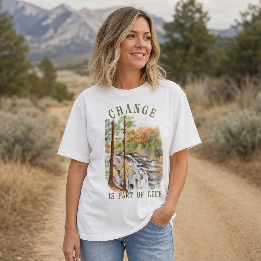 Woman wearing a white Comfort Colors 1717 t-shirt featuring a watercolor forest waterfall and pine tree design with vintage text 'Change is part of life', standing on a scenic mountain trail outdoors, by Crossroads Dandelion